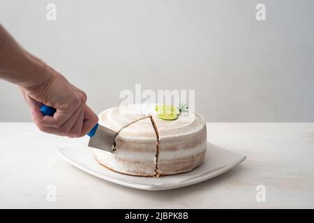 La mano di un uomo taglia una torta di compleanno Vegan con mandorle, cocco e limone base con un coltello grande su un tavolo bianco e uno sfondo bianco Foto Stock