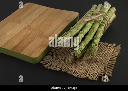 Mazzo di asparagi verdi legato con filo di iuta vicino al tagliere su sfondo nero con spazio copia. Cottura degli asparagi e concep di verdure primaverili Foto Stock