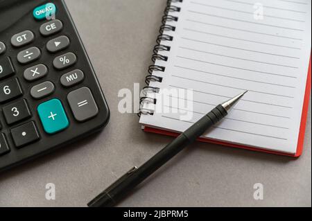 Calcolatrice, penna, blocco note giacciono su una superficie grigia. Aprire il notebook caricato a molla con fogli vuoti. Primo piano. Foto Stock