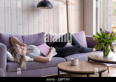 Problemi di sonno. Bella bionda in T-shirt rosa e jeans si addormentarono a casa sul divano con il computer portatile in mano. Lavora a casa online. Fine settimana. Lavoro eccessivo Foto Stock