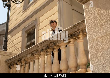 Ritratto di donna con lunghi capelli lisci con occhiali da sole neri, maglione bianco, in piedi al balaustra di marmo beige. Foto Stock