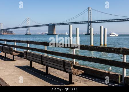 Panca vicino recinzione sul bordo della spiaggia di fronte al mare con vista di San Francisco - Oakland Bay Bridge della giornata di sole in California, Stati Uniti. Foto Stock