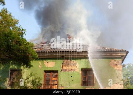 Un getto d'acqua proveniente da un idrante antincendio spegne il tetto che brucia e fuma di una casa. Foto Stock