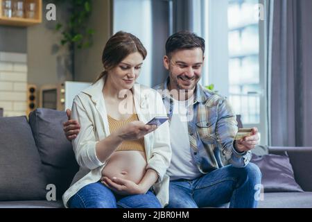 Giovane coppia sposata marito e donna incinta seduta insieme sul divano e abbracciare utilizzando il telefono per lo shopping online nel negozio online, hol Foto Stock