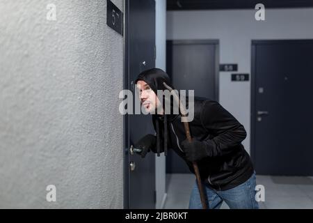 Il ladro cerca di rompere la serratura della porta nell'appartamento, vestito di nero Foto Stock