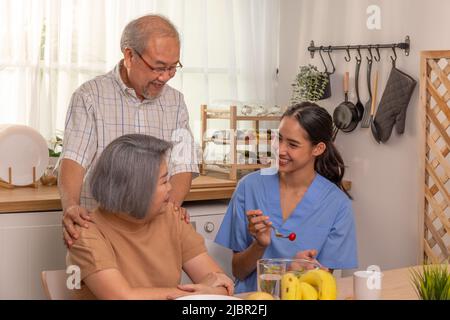 Una giovane infermiera asiatica fornisce cibo per un uomo anziano al servizio di casa di cura in pensione. Un operatore sanitario sostiene gli anziani in cucina durante la prima colazione. Foto Stock