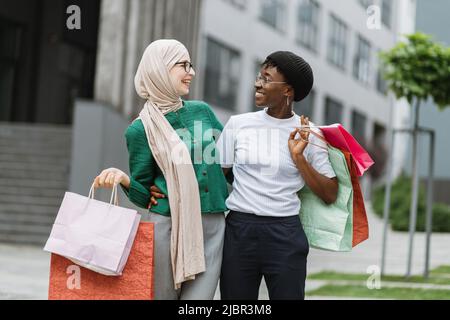 Due felici ragazze multietniche soddisfatti, camminando lungo la strada della città vecchia dopo lo shopping, portando le borse colorate nelle loro mani, sorridendo alla macchina fotografica. Shopping in città, stile di vita urbano. Foto Stock