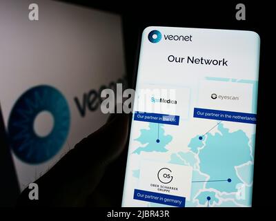 Persona che tiene il cellulare con pagina web della rete clinica oftalmologica Veonet GmbH sullo schermo di fronte al logo. Concentrarsi sul centro del display del telefono. Foto Stock