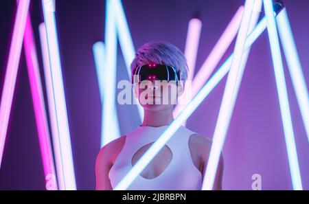 3D rendering di giovane donna elegante con capelli biondi corti in futuristici occhiali VR che esplorano la realtà virtuale in piedi tra bastoni al neon illuminati Foto Stock