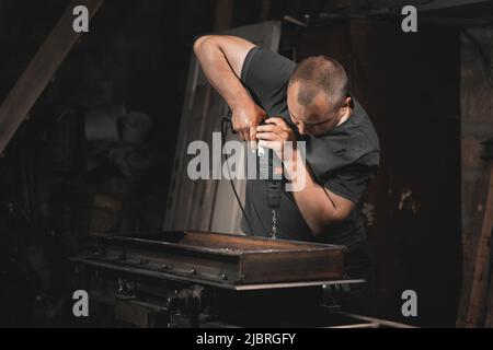 Un uomo perfora il metallo con un trapano a mano nella sua officina domestica. Realizzare un prodotto in metallo con le proprie mani Foto Stock