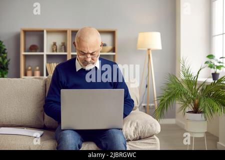 Uomo anziano in occhiali seduti sul divano a casa e lavorando sul suo computer portatile Foto Stock