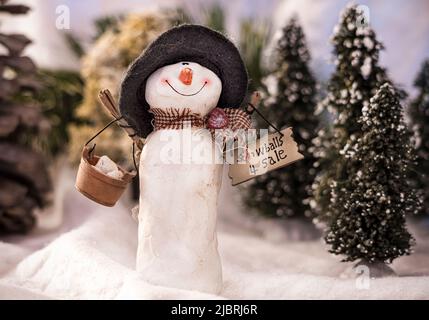 Un paesaggio innevato in miniatura di alberi sempreverdi coperti di neve con un pupazzo di neve che sorride mentre tiene un cesto di palle di neve e palle di neve per la vendita segno Foto Stock