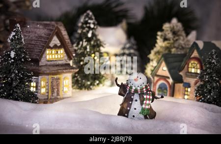 Un villaggio illuminato in miniatura di notte coperto di neve con un pupazzo di neve accogliente di fronte Foto Stock