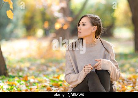Giovane donna di razza mista pensiva seduta sull'erba nel parco all'inizio dell'autunno Foto Stock