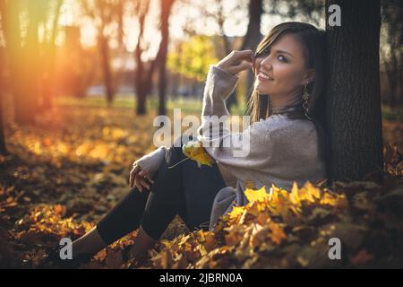 Giovane felice misto gara donna seduta vicino albero in autunno parco al tramonto guardando la macchina fotografica Foto Stock