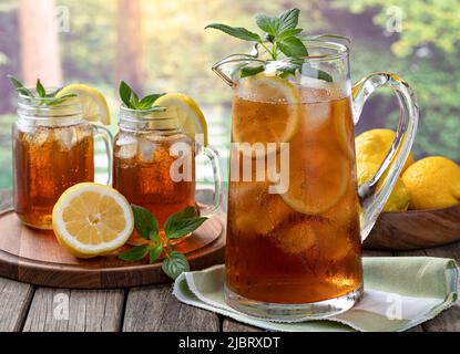Caraffa di tè freddo ghiacciato con menta, fette di limone e ghiaccio con due bicchieri di tè su un tavolo di legno e sfondo rurale estivo Foto Stock