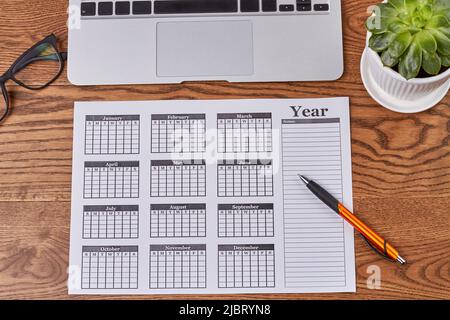 Vista dall'alto calendario dell'anno con penna e pianta su scrivania in legno. Calendario aziendale piatto e altri accessori. Foto Stock