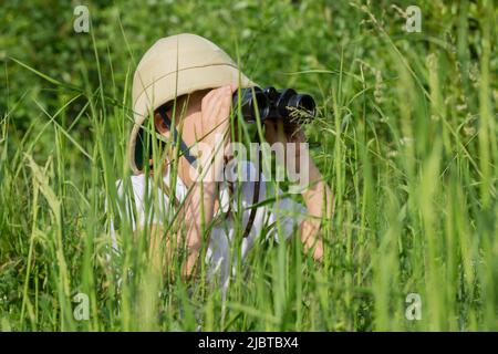 Ragazza che indossa il casco di pith nascosto in erba guardando attraverso binocoli osservare la natura estiva. Concetto di scoperta e avventure Foto Stock