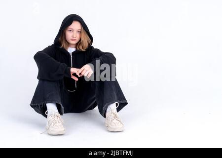 Una adolescente caucasica in una felpa nera con cappuccio è seduta sul pavimento su sfondo bianco. Foto studio. Adolescenza difficile transizione Foto Stock