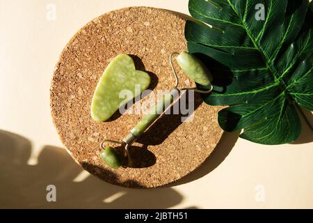 Raschietto Jade Gua sha e massaggiatore a rullo sul supporto rotondo in sughero con una foglia di monstera. Luce dura, ombre, il concetto di auto-cura. Cura del viso. Zer Foto Stock