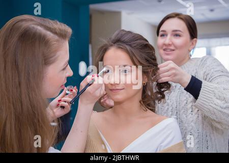 Due maestri con un sorriso fanno trucco e capelli a una ragazza nel salone. Make-up artista e parrucchiere creare un'immagine di una giovane donna. Business Concept - salone di bellezza, cura del viso e dei capelli. Foto Stock