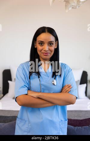 Ritratto di fidata fisioterapista biraciale femminile con le braccia incrociate in piedi a casa, copia spazio Foto Stock