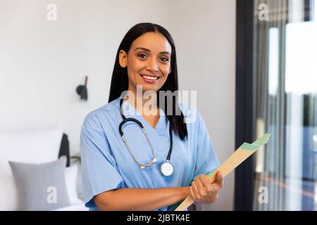 Ritratto di fisioterapista femminile felice biraciale con rapporto e stetoscopio in piedi a casa Foto Stock