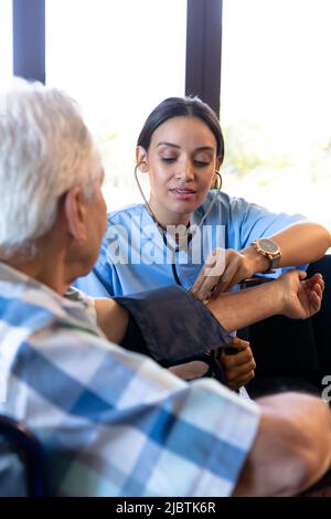 Medico biracale femminile che esamina la pressione sanguigna dell'uomo maggiore caucasico con indicatore e stetoscopio Foto Stock