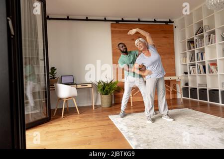 Fisioterapista maschile afroamericano che aiuta la donna anziana caucasica a svolgere l'esercizio di stretching Foto Stock