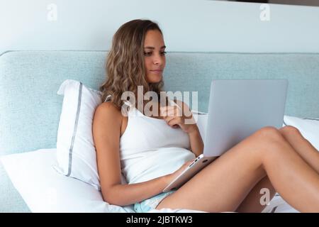 Giovane donna caucasica che usa un computer portatile sdraiato sul letto di casa Foto Stock