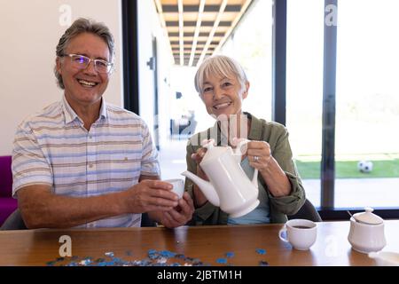 Ritratto di sorridente donna anziana asiatica che versa caffè in tazza tenuto da uomo anziano caucasico a tavola Foto Stock