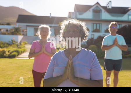 Gli anziani multirazziale meditating mentre si levano in piedi contro la casa di pensionamento in iarda il giorno di sole Foto Stock