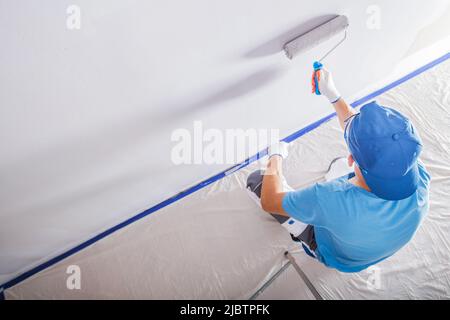Abbigliamento maschile con T-shirt blu e cappellino da baseball blu durante i lavori di ristrutturazione verniciatura delle pareti in una stanza bianca con un rullo di vernice. Foto Stock