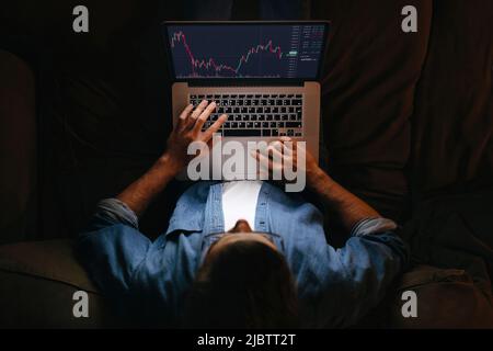 Giovane uomo di affari analista di commerciante che guarda il monitor del laptop sul divano, intermediario dell'investitore che analizza gli indici, grafico finanziario che commercia i dati di investimento in linea sul grafico del mercato azionario di criptovaluta. Vista dall'alto Foto Stock