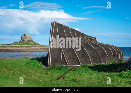 Coast UK, vista di una barca da pesca capovolta che serve come capanna di stoccaggio su Holy Island con il castello di Lindisfarne in lontananza, Northumberland costa, Regno Unito Foto Stock