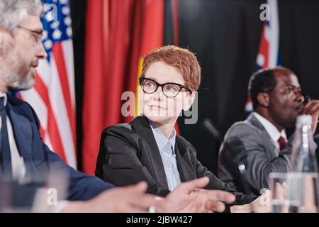 Giovane donna d'affari in occhiali che ascolta la sua collega mentre siedono alla conferenza Foto Stock