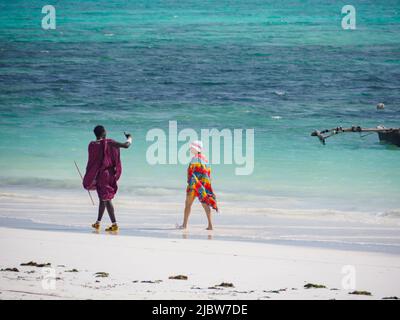 Zanzibar, Tanzania - Gennaio 2021: Guerriero africano Maasai in abito tradizionale con donna caucasica che cammina su una spiaggia sabbiosa sullo sfondo del blu Foto Stock