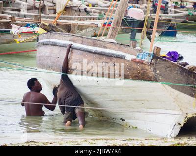 Kizimkazi, Zanzibar - Jan, 2021: Un pescatore pulisce una tradizionale barca in sambuco di legno in piedi sulla riva dell'oceano a bassa marea. Zanzibar, Tanzania, Africa Foto Stock