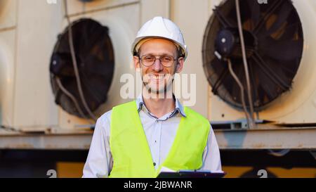 Ritratto di ingegnere sorridente in casco facendo diagnosi di un impianto di climatizzazione industriale con una clipboard. Foto Stock