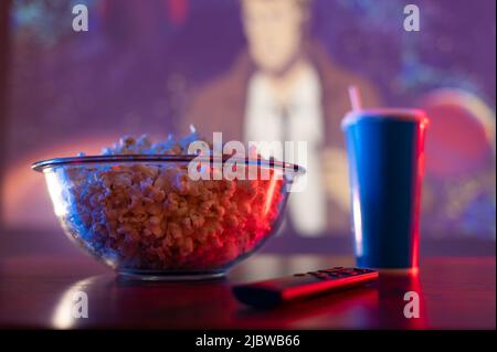 Un set classico per il relax - una ciotola di vetro con popcorn e una bevanda analcolica in un bicchiere, un telecomando TV. Lo sfondo è un film televisivo. Ricreazione, r Foto Stock