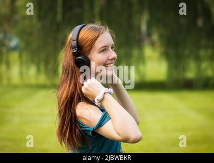 Ragazza adolescente con lunghi capelli rossi sorridenti e indossando cuffie in un campo verde con un albero di salice e prato verde sullo sfondo durante l'estate Foto Stock