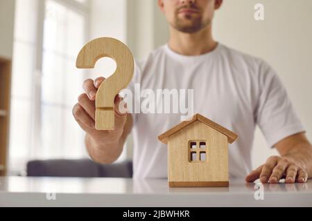 Confusione giovane uomo che è esitante circa l'acquisto della sua casa mostra legno punto di domanda.