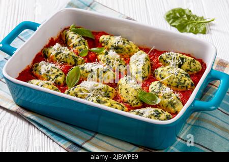 Malfatti, gnocchi di ricotta di spinaci italiani in salsa di pomodoro con erbe aromatiche e parmigiano grattugiato in piatto di cottura in ceramica su tavola bianca Foto Stock