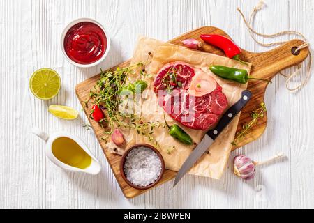 osso buco crudo, bistecca di vitello con marrowbone su tavola di legno con pepe in grani, timo fresco, peperoncino, aglio e lime su tavola di legno bianco, orizzonte Foto Stock