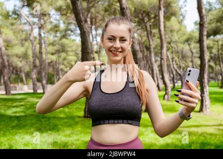 Donna allegra bruna che indossa abiti sportivi sul parco cittadino, all'aperto tenendo un telefono e puntando il dito al telefono con il viso sorridente. Sta cercando Foto Stock