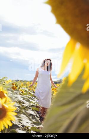 La ragazza cammina in un campo di girasole in una calda giornata di sole. Una donna cammina tra i fiori gialli e dorati. I semi sono cresciuti per il consumo umano o per la produzione di petrolio. Messa a fuoco selettiva soft Foto Stock