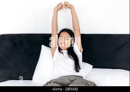 Felice ragazza bruna asiatica si sveglia, si allunga al mattino il fine settimana mentre si siede sul letto in una camera da letto, solleva le mani con gli occhi chiusi, sorridendo felicemente Foto Stock