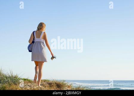 Vista posteriore della giovane donna che porta la sua borsa e scarpe camminando lungo una costa sabbiosa in spiaggia Foto Stock