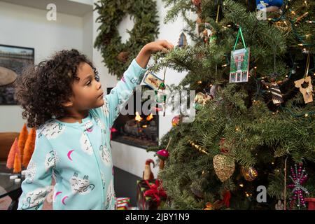 Bambino che raggiunge fino ad appendere un ornamento sull'albero illuminato di Natale nella sua casa Foto Stock