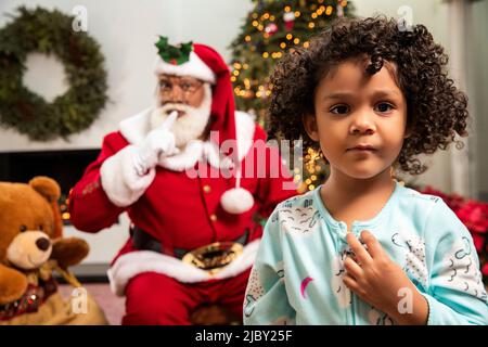 Ragazza che guarda in macchina fotografica con suo nonno vestito come Babbo Natale sullo sfondo. Divertimento di Natale. Foto Stock